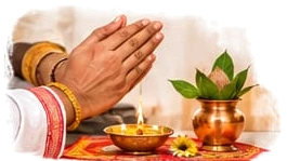 Hands folded in prayer for the sankalpa, beside a lit diya and a copper kalash with mango leaves
